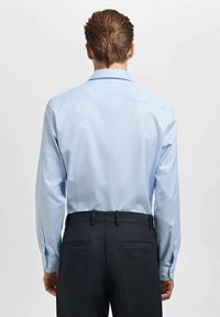 Svetlomodrá košeľa s dlhými rukávmi a jemnou textúrou, s klasickým golierom a manžetami na gombíky, v kombinácii s tmavými nohavicami.