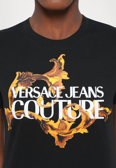 T-shirt en coton noir avec un imprimé orné vibrant en orange et jaune, portant le texte "VERSACE JEANS COUTURE" en lettres blanches épaisses.