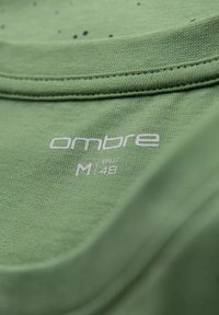 Grøn bomuldsstof med en glat tekstur. Har hvidt trykt tekst "ombre M 48" på kraven. Enkel og ren design.
