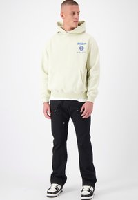 Lichtbeige Hoodie met een voorzak, logo in het blauw; gecombineerd met zwarte broek met zilveren hardware en sneakers met witte en zwarte accenten.