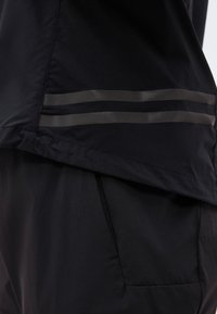 Gros plan sur un tissu noir avec deux bandes réfléchissantes horizontales près de l'ourlet, superposé sur un pantalon noir avec des coutures de poche visibles.