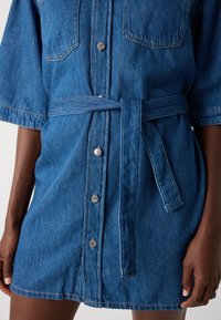 Denim srajca obleka v modri barvi s kratkimi rokavi, gumbnim zapiranjem spredaj, dvema žepoma na prsih in pasom iz istega blaga, ki poudari pas.
