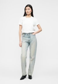 Νεαρή γυναίκα που φοράει απλό λευκό μπλουζάκι, ανοιχτό μπλε ψηλόμεσο τζιν και μαύρες μποτάκια στον αστράγαλο, στέκεται με το ένα χέρι στην τσέπη.