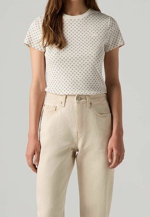 Mujer con una blusa blanca de manga corta con estampado, metida en unos vaqueros de tiro alto color crema, sobre un fondo liso.