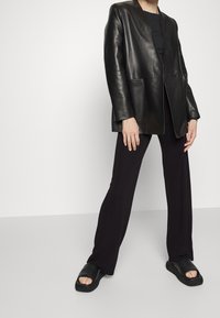 Černý kožený blejzr s mírným leskem, přiléhavý střih a klopové límce, v kombinaci s černými širokými kalhotami a černými skluzovkami.