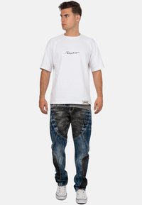 T-shirt blanc à manches courtes avec un texte graphique noir, associé à un jean bleu avec un motif camouflage et des accents de boutons sur le côté. Baskets blanches.