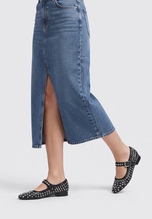 Persona con una falda midi vaquera azul con abertura frontal y zapatos Mary Jane negros con tachuelas y correa con hebilla.