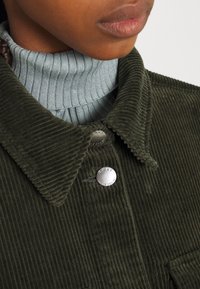 Olivově zelená kordurová bunda s límečkem, stříbrnými cvočky a světle modrým žebrovaným rolákem pod ní.