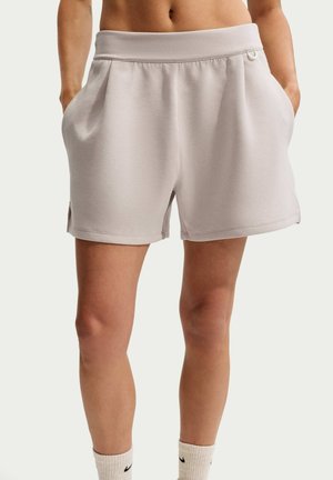 Short ample beige clair avec poches latérales porté par une personne debout, montré de la taille à mi-cuisse, sur un fond uni.