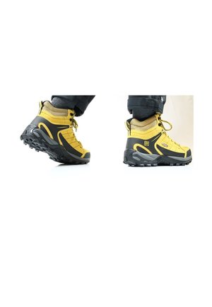 Botas de senderismo amarillas y negras usadas con pantalones oscuros, mostrando vistas laterales y anguladas sobre un fondo blanco.