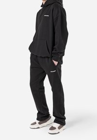 Svart hoodie med framficka och logotyp, kombinerad med svarta sweatpants med logotyp, bärs med vita och grå sneakers.