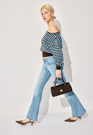 Jeune femme marchant, vêtue d'un haut rayé à épaules dénudées, d'un jean bleu clair, de petits talons marron, tenant un petit sac à main marron orné d'un foulard.