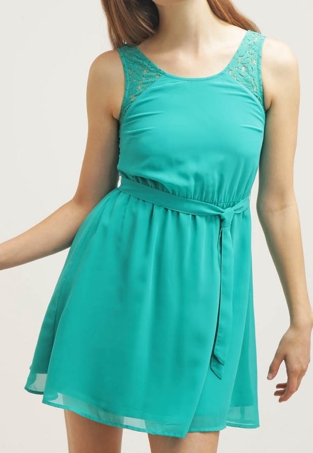 Femme portant une robe sans manches de couleur teal avec des panneaux en dentelle aux épaules et une ceinture nouée à la taille, se tenant devant un fond clair et uni.