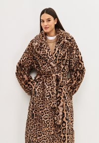 Abrigo de pelo sintético con estampado de leopardo, con un cuello ancho, cinturón en la cintura y dos bolsillos laterales. Textura suave con patrones en marrón oscuro y negro sobre fondo beige.