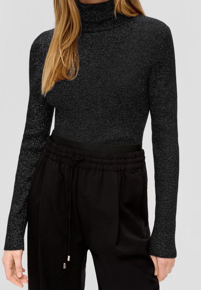Femme portant un pull à col roulé noir ajusté et pailleté ainsi qu'un pantalon noir ample avec cordon, montrée du cou aux hanches.