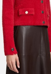 Czerwona, puszysta kurtka z srebrnymi guzikami w zestawieniu z ciemnobrązową skórzaną spódnicą o wysokim stanie, ręka oparta na boku, neutralne tło.