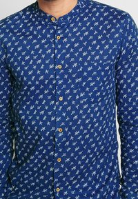 Blå knäppt skjorta med ett blommönster bestående av små vita blad, rundad krage och lätt textur. Träknappar ger detaljer.