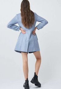 Donna con lunghi capelli mossi, che indossa un cappotto azzurro chiaro, pantaloni corti neri e stivali neri spessi, in piedi con le mani posate sulla parte bassa della schiena.
