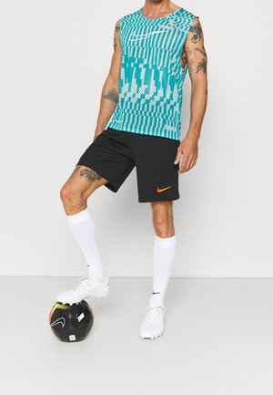 Haut sans manches turquoise Nike avec motif, short noir avec logo orange, chaussettes blanches et chaussures blanches, debout sur un ballon de football noir.