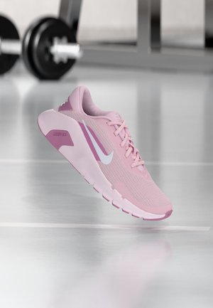 Roze Nike hardloopschoen met witte swoosh en flexibele zool zwevend in sportschoolomgeving met wazige gewichten op de achtergrond.