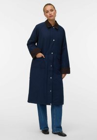 Cappotto lungo blu navy con colletto e polsini marroni, bottoni a pressione, tasche laterali e taglio dritto. Il tessuto appare texturizzato e resistente.