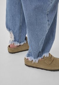 Lyseblå denimjeans med frynsede kanter, kombineret med brune mokkasin-lignende sko i ruskind, der udstråler en blød tekstur og et afslappet design.