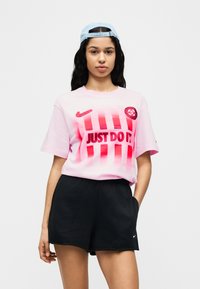 Ροζ κοντομάνικη μπλούζα με μεγάλο μαύρο σύνθημα «JUST DO IT» και κόκκινες ρίγες. Συνδυάζεται με μαύρο σορτς, διαθέτει λογότυπα Nike.