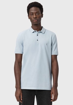 Mann trägt ein hellblaues Polohemd mit kurzen Ärmeln und schwarze Hose, blickt nach vorne vor einem einfachen grauen Hintergrund.