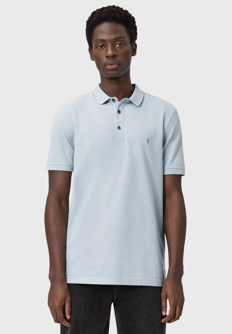 Mężczyzna ubrany w jasnoniebieską koszulkę polo z krótkim rękawem i czarne spodnie, stojący przodem na jednolitym szarym tle.