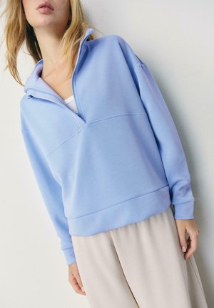 Femme portant un sweat-shirt bleu clair à demi-fermeture éclair par-dessus un haut blanc et un pantalon large beige, debout devant un fond clair uni.