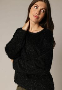 Jonge vrouw met lang bruin haar die een zwarte fluffy trui draagt, iets omhoog kijkend met haar hand op haar nek.