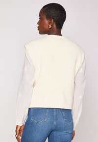 Crème-kleurige gebreide vest met een textuurpatroon, schoudernaden en een geribde zoom, gedragen over een lichtbeige langemouwen shirt met bloemaccenten.