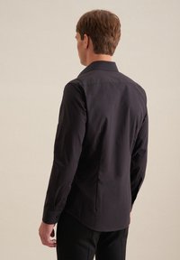 Uomo che indossa una camicia nera a maniche lunghe ben aderente e pantaloni neri, rivolto di spalle contro uno sfondo beige uniforme.