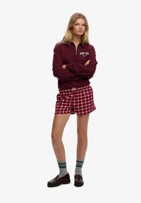 Bordowa bluza z zamkiem i napisem "NEW YORK" oraz bordowe spodenki w kratę. Model nosi szare skarpety z zielonymi paskami i ciemne mokasyny.