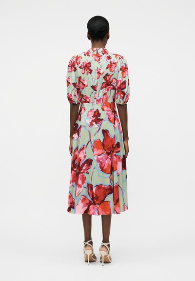 Γυναίκα που στέκεται πλάτη προς τα εμπρός, φοράει πράσινο μίντι φόρεμα με μεγάλα κόκκινα και ροζ λουλουδένια σχέδια και φουσκωτά μανίκια μέχρι τον αγκώνα.