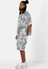 Camicia a maniche corte con bottoni e pantaloni corti coordinati con stampa tropicale bianca e nera. Indossato con scarpe da ginnastica bianche e calze alla caviglia.