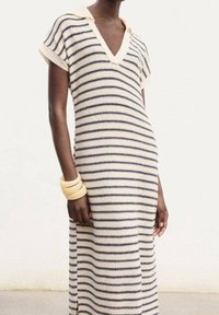 Personne à la peau foncée portant une robe en maille à manches courtes crème et bleu marine rayée avec un col, ainsi que des bracelets rigides beiges au poignet gauche.