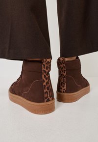 Zapatillas altas marrones con detalles de estampado de leopardo, material de ante, lados texturizados y suela de goma. Detalle de costura lateral visible.