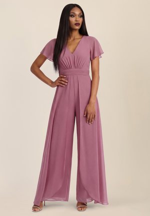 Frau trägt einen mauvefarbenen Jumpsuit mit weitem Bein, flatternden Ärmeln und V-Ausschnitt, steht mit einer Hand in der Hüfte vor einem einfarbigen Hintergrund.