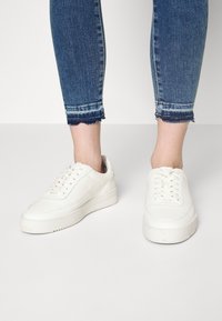 Zapatillas de cuero blancas con un acabado suave, punta redondeada y suela de goma gruesa. Se pueden ver los cordones y detalles de costura sutiles.