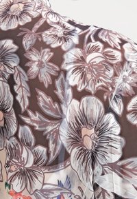 Blouse semi-transparente à motif floral avec un fond marron foncé et des motifs floraux rose clair et bleu. Le tissu a une texture légère.