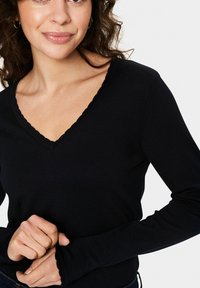 Juodas ilgomis rankovėmis viršus su V formos iškirpte ir banguota apdaila, pagamintas iš tekstūruoto raukšlėto audinio. Prigludusi forma su aptemptomis rankovėmis.