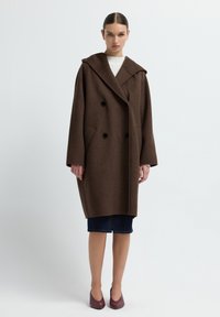 Brauner Wollmantel mit doppelreihigem Schnitt, großen Revers und Taschen. Getragen über einem weißen Pullover und einem Jeansrock, kombiniert mit burgunderfarbenen Schuhen.