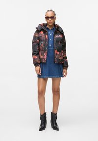 Casaco preto acolchoado com estampa floral colorida, com fecho de correr à frente e capuz, usado sobre um vestido de ganga. A modelo usa óculos de sol e botins pretos.