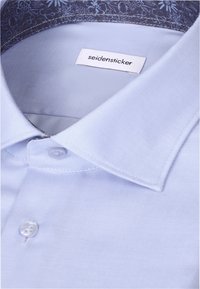 Col de chemise bleu clair avec motif floral à l'intérieur du col et étiquette de marque "seidensticker" visible.