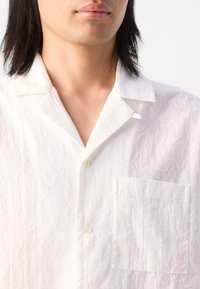 White short-sleeve shirt with a textured pattern, featuring a notch collar, subtle stitching, and a patch pocket on the left side.
