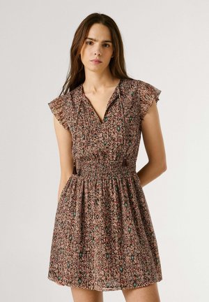 Jonge vrouw die een halflange, bedrukte jurk draagt met een geplooide taille en kwastjes, staand tegen een effen lichte achtergrond.