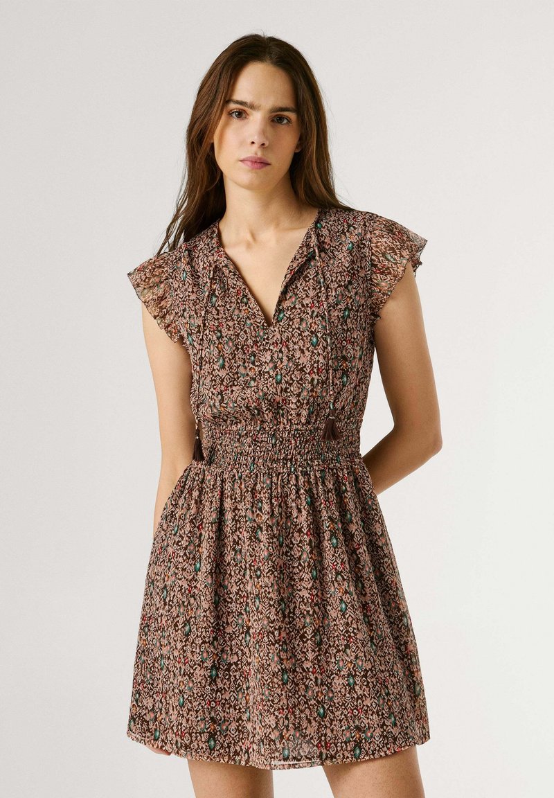 Mlada ženska, oblečena v kratko obleko z vzorcem, z nabrano pasom in trakovi z resicami, stoji pred enobarvno svetlo ozadje.