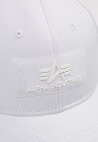 Bijela bejzbol kapa s teksturiranim prednjim dijelom i utisnutim logotipom i imenom brenda "Alpha Industries". Glatka tkanina s prošivenim detaljima.