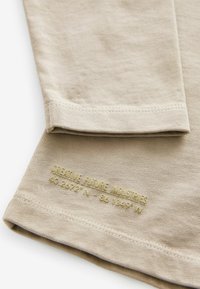 Beige pitkähihaisessa paidassa on pehmeä kangas, jossa on näkyvä helma ja kohotulostettu kultainen teksti alaosassa.